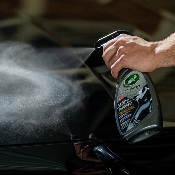 Polishing a black car. Hand sprays Turtle Wax Hybrid Solutions Ceramic Acrylic Black Wax on a dark, glossy auto surface.