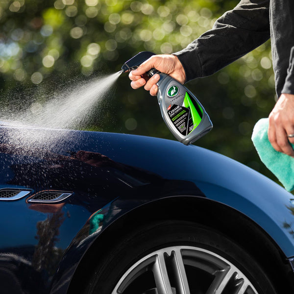 Turtle Wax Hybrid Solutions ceramic car wax spray being applied to a blue vehicle's hood by hand, with a microfiber towel visible.